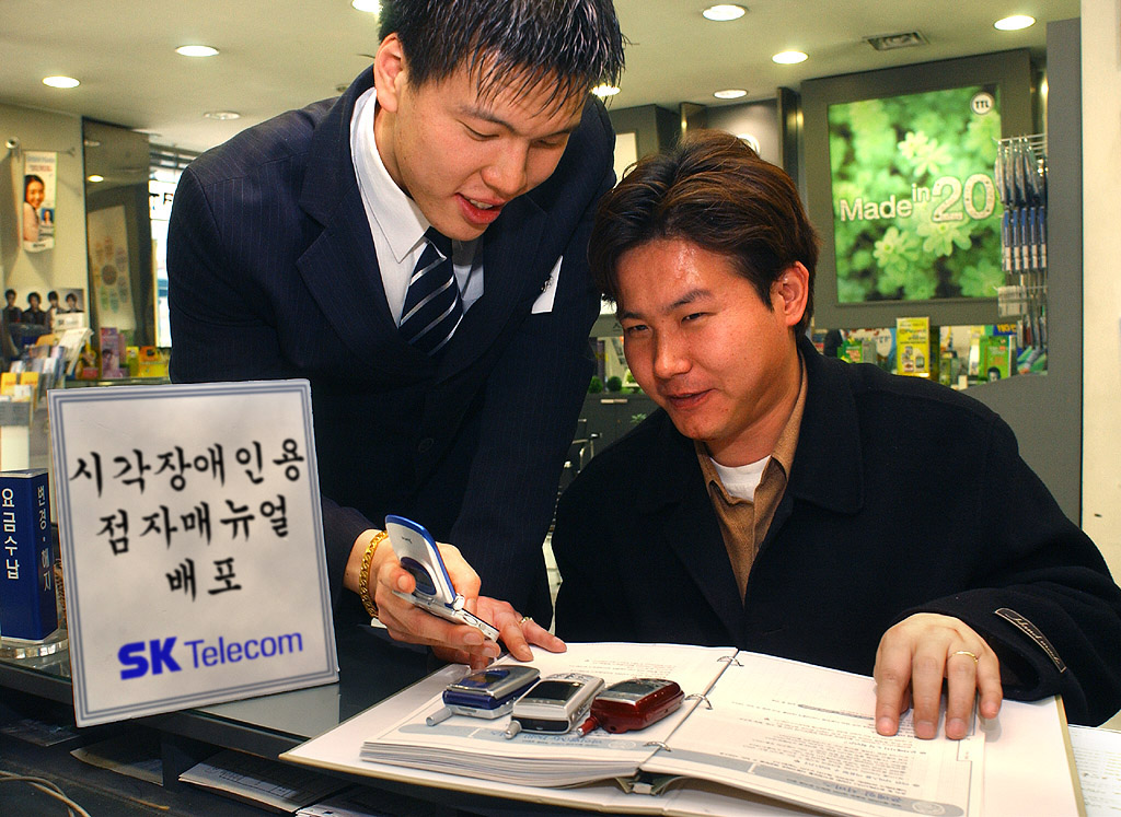 Visually impaired customers are shown in an SK Telecom sales outlet, learning how to use cellular services through the upgraded Braille version of the SPEED 011 User`s Guide